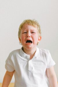 A young child crying, wearing a white shirt, showing strong emotions indoors.